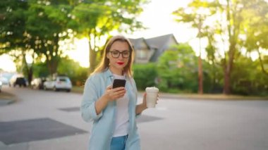 Akıllı telefonlu ve elinde kahve olan mutlu bir kadın yaz akşamı mahallede dolaşıyor..