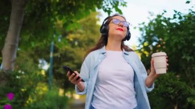 Gözlüklü ve kulaklıklı mutlu kadın elinde kahve ve akıllı telefonuyla çiçek açan bahçede ya da yaz günü yeşil sokakta yürüyor..