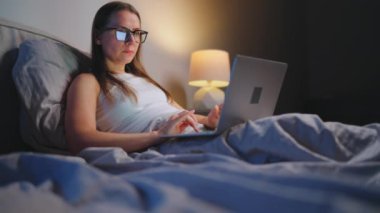 Gözlüklü kadın geceleri yatakta yatarken dizüstü bilgisayarda çalışıyor. İnternet bağımlısı veya uykusuzluk konsepti. 