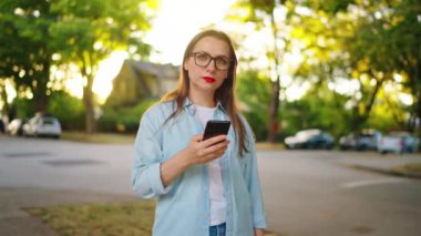 Mutlu kadın güneşli bir yaz gününde yeşil caddede dikilirken akıllı telefon kullanıyor. Yavaş çekim