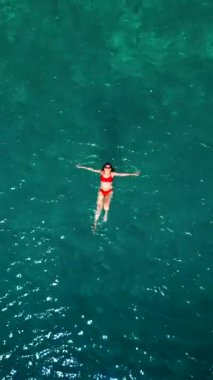 Kırmızı mayo giymiş bir kadının havadan görünüşü, kristal berrak deniz sularında rahatlama somutlaştırıcı bir şekilde zahmetsizce sırt üstü yüzüyor. Yaz tatilleri. Dikey video