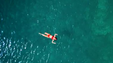 Kırmızı mayo giymiş bir kadının havadan görünüşü, kristal berrak deniz sularında rahatlama somutlaştırıcı bir şekilde zahmetsizce sırt üstü yüzüyor. Yaz tatilleri.