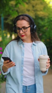 Gözlüklü, kulaklıklı, elinde kahve ve akıllı telefonuyla yaz günü yeşil bir sokakta yürüyen mutlu bir kadın..