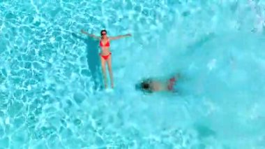 Çift havuzda rahatlar. Kadın sırtüstü yatarken, bir erkeğin havuza dalıp yüzdüğü tepeden aşağı manzara..