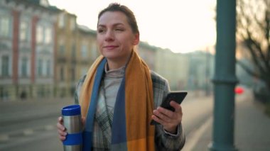 Beyaz ceketli bir iş kadını, sabahın erken saatlerinde şehirde dolaşıyor, kahve içiyor ve akıllı telefon kullanıyor, ağır çekimde. İletişim, iş günü, yoğun yaşam konsepti.