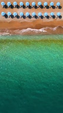 Mavi güneş şemsiyeleri ve turkuaz deniz suyuyla cennet gibi kumlu sahilin yukarıdan görünüşü. Dikey video