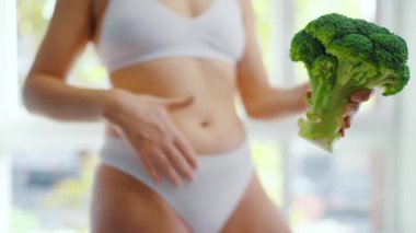Bir kadın elinde bir brokoli tutuyor. Karnını okşuyor ve baş parmağını kaldırıyor. Sağlıklı beslenme ve sindirim kavramı