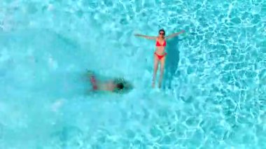 Çift havuzda rahatlar. Kadın sırtüstü yatarken, bir erkeğin havuza dalıp yüzdüğü tepeden aşağı manzara..