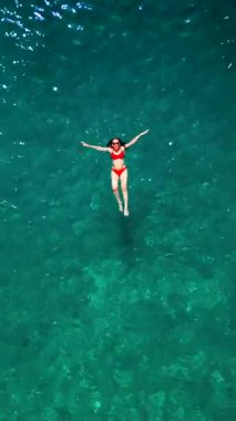 Kırmızı mayo giymiş bir kadının havadan görünüşü, kristal berrak deniz sularında rahatlama somutlaştırıcı bir şekilde zahmetsizce sırt üstü yüzüyor. Yaz tatilleri. Dikey video