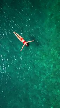 Kırmızı mayo giymiş bir kadının hava görüntüsü, kristal berrak deniz sularında rahatlama somutlaştırıcı bir şekilde sırtüstü uzanır. Yaz tatilleri. Dikey video