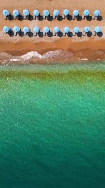 Mavi güneş şemsiyeleri ve turkuaz deniz suyuyla cennet gibi kumlu sahilin yukarıdan görünüşü. Dikey video