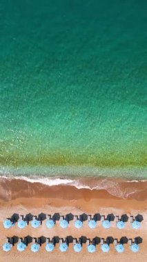 Mavi güneş şemsiyeleri ve turkuaz deniz suyuyla cennet gibi kumlu sahilin yukarıdan görünüşü. Dikey video