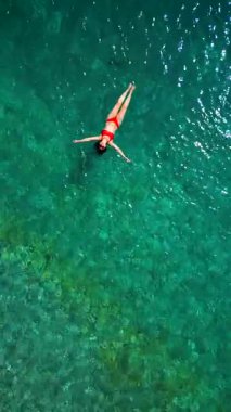 Kırmızı mayo giymiş bir kadının hava görüntüsü, kristal berrak deniz sularında rahatlama somutlaştırıcı bir şekilde sırtüstü uzanır. Yaz tatilleri. Dikey video