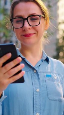 Gözlüklü beyaz kadın sokakta duruyor, akıllı telefon kullanıyor ve kahve içiyor. Yörünge atışı. Arka planda gökdelenler var. İletişim, iş günü, yoğun yaşam konsepti.