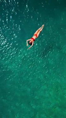 Kırmızı mayo giymiş bir kadının hava görüntüsü, kristal berrak deniz sularında rahatlama somutlaştırıcı bir şekilde sırtüstü uzanır. Yaz tatilleri. Dikey video
