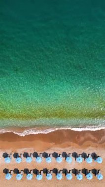 Mavi güneş şemsiyeleri ve turkuaz deniz suyuyla cennet gibi kumlu sahilin yukarıdan görünüşü. Dikey video