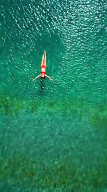 Kırmızı mayo giymiş bir kadının hava görüntüsü, kristal berrak deniz sularında rahatlama somutlaştırıcı bir şekilde sırtüstü uzanır. Yaz tatilleri. Dikey video