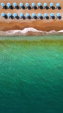 Mavi güneş şemsiyeleri ve turkuaz deniz suyuyla cennet gibi kumlu sahilin yukarıdan görünüşü. Dikey video