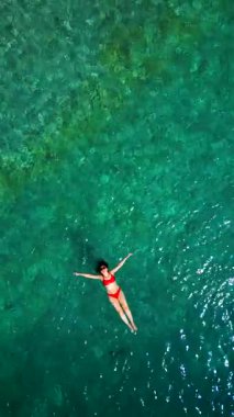 Kırmızı mayo giymiş bir kadının hava görüntüsü, kristal berrak deniz sularında rahatlama somutlaştırıcı bir şekilde sırtüstü uzanır. Yaz tatilleri. Dikey video