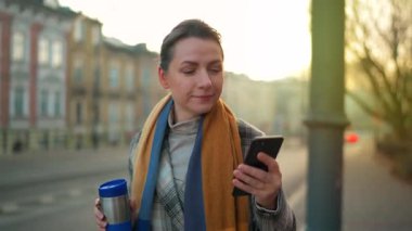 Beyaz ceketli bir iş kadını, sabahın erken saatlerinde şehirde dolaşıyor, kahve içiyor ve akıllı telefon kullanıyor. İletişim, iş günü, yoğun yaşam konsepti.