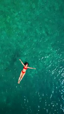Kırmızı mayo giymiş bir kadının hava görüntüsü, kristal berrak deniz sularında rahatlama somutlaştırıcı bir şekilde sırtüstü uzanır. Yaz tatilleri. Dikey video