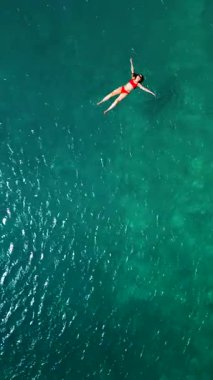 Kırmızı mayo giymiş bir kadının hava görüntüsü, kristal berrak deniz sularında rahatlama somutlaştırıcı bir şekilde sırtüstü uzanır. Yaz tatilleri. Dikey video