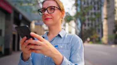 Gözlüklü beyaz kadın şehrin caddesinde dikiliyor ve akıllı telefon kullanıyor. Arka planda gökdelenler var. İletişim, iş günü, yoğun yaşam konsepti.