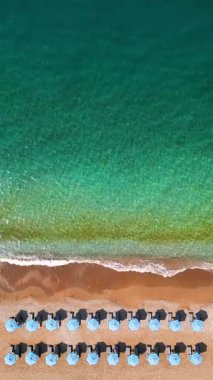 Mavi güneş şemsiyeleri ve turkuaz deniz suyuyla cennet gibi kumlu sahilin yukarıdan görünüşü. Dikey video