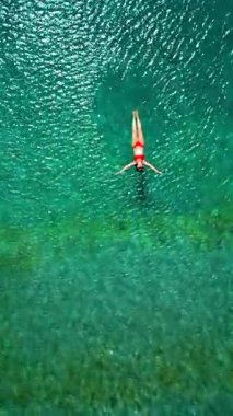 Kırmızı mayo giymiş bir kadının hava görüntüsü, kristal berrak deniz sularında rahatlama somutlaştırıcı bir şekilde sırtüstü uzanır. Yaz tatilleri. Dikey video
