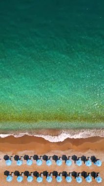 Mavi güneş şemsiyeleri ve turkuaz deniz suyuyla cennet gibi kumlu sahilin yukarıdan görünüşü. Dikey video