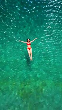 Kırmızı mayo giymiş bir kadının hava görüntüsü, kristal berrak deniz sularında rahatlama somutlaştırıcı bir şekilde sırtüstü uzanır. Yaz tatilleri. Dikey video