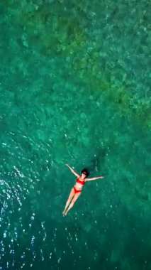 Kırmızı mayo giymiş bir kadının hava görüntüsü, kristal berrak deniz sularında rahatlama somutlaştırıcı bir şekilde sırtüstü uzanır. Yaz tatilleri. Dikey video
