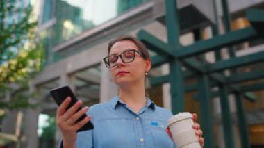 Gözlüklü beyaz kadın sokakta duruyor, akıllı telefon kullanıyor ve kahve içiyor. Yörünge atışı. Arka planda gökdelenler var. İletişim, iş günü, yoğun yaşam konsepti.