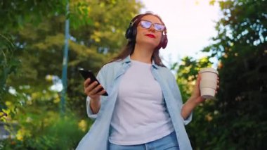 Gözlüklü, kulaklıklı, elinde kahve ve akıllı telefonuyla yaz günü yeşil bir sokakta yürüyen mutlu bir kadın..
