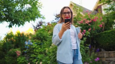 Dizüstü bilgisayarı ve akıllı telefonu olan mutlu bir kadın çiçek açan bahçede ya da işten sonra yeşil sokakta duruyor..