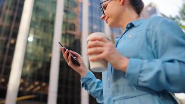 Gözlüklü beyaz kadın sokakta duruyor, akıllı telefon kullanıyor ve kahve içiyor. Yörünge atışı. Arka planda gökdelenler var. İletişim, iş günü, yoğun yaşam konsepti.