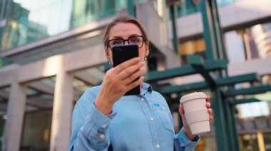 Gözlüklü beyaz kadın sokakta duruyor, akıllı telefon kullanıyor ve kahve içiyor. Yörünge atışı. Arka planda gökdelenler var. İletişim, iş günü, yoğun yaşam konsepti.