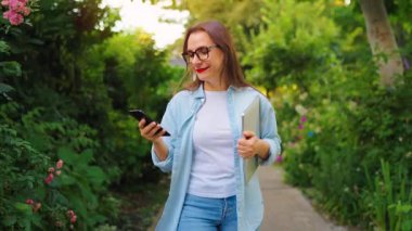 Dizüstü bilgisayarı ve akıllı telefonu olan mutlu bir kadın çiçek açan bahçede ya da iş gününden sonra yeşil sokakta yürüyor. Yavaş çekim