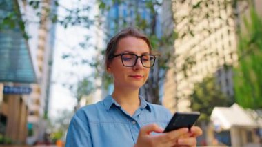 Gözlüklü beyaz kadın şehirde dolaşıyor ve akıllı telefon kullanıyor, ağır çekimde. Arka planda gökdelenler var. İletişim, iş günü, yoğun yaşam konsepti.