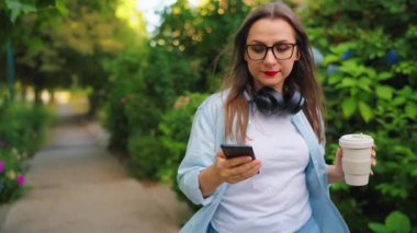 Gözlüklü, kulaklıklı, elinde kahve ve akıllı telefonuyla yaz günü yeşil bir sokakta yürüyen mutlu bir kadın..