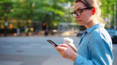 Gözlüklü beyaz kadın şehirde dolaşıyor, akıllı telefon kullanıyor ve kahve içiyor, ağır çekimde. Arka planda gökdelenler var. İletişim, iş günü, yoğun yaşam konsepti.