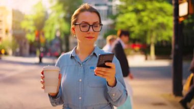 Gözlüklü beyaz kadın sokakta duruyor, akıllı telefon kullanıyor ve kahve içiyor, ağır çekimde. Yörünge atışı. Arka planda gökdelenler var. İletişim, iş günü, yoğun yaşam konsepti.