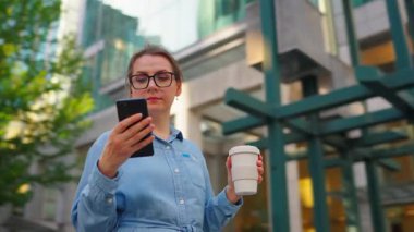Gözlüklü beyaz kadın sokakta duruyor, akıllı telefon kullanıyor ve kahve içiyor. Yörünge atışı. Arka planda gökdelenler var. İletişim, iş günü, yoğun yaşam konsepti.