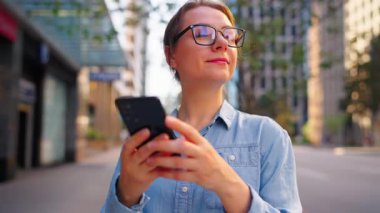 Gözlüklü beyaz kadın şehrin caddesinde dikiliyor ve akıllı telefon kullanıyor. Arka planda gökdelenler var. İletişim, iş günü, yoğun yaşam konsepti.