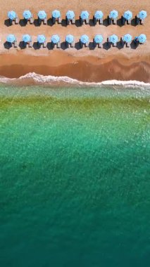 Mavi güneş şemsiyeleri ve turkuaz deniz suyuyla cennet gibi kumlu sahilin yukarıdan görünüşü. Dikey video