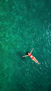 Kırmızı mayo giymiş bir kadının hava görüntüsü, kristal berrak deniz sularında rahatlama somutlaştırıcı bir şekilde sırtüstü uzanır. Yaz tatilleri. Dikey video