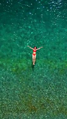 Kırmızı mayo giymiş bir kadının hava görüntüsü, kristal berrak deniz sularında rahatlama somutlaştırıcı bir şekilde sırtüstü uzanır. Yaz tatilleri. Dikey video