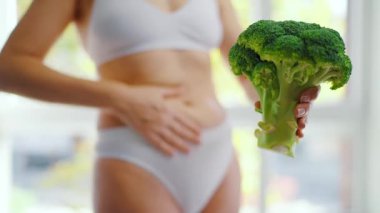 Bir kadın elinde bir brokoli tutuyor. Karnını okşuyor ve baş parmağını kaldırıyor. Sağlıklı beslenme ve sindirim kavramı