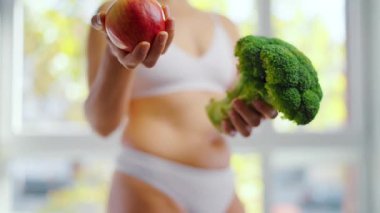 Bir kadın elinde brokoli ve elmayla dans ediyor. Hangisinin daha iyi olduğuna o karar verir. Sağlıklı beslenme kavramı.