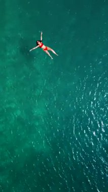 Kırmızı mayo giymiş bir kadının hava görüntüsü, kristal berrak deniz sularında rahatlama somutlaştırıcı bir şekilde sırtüstü uzanır. Yaz tatilleri. Dikey video